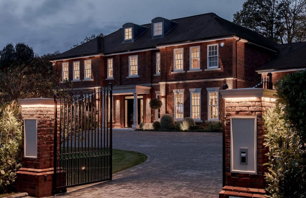 Front view of a 10,000 sq ft new build property in Kingswood, Surrey, showcasing red brick architecture, wrought iron gates and warm exterior lighting