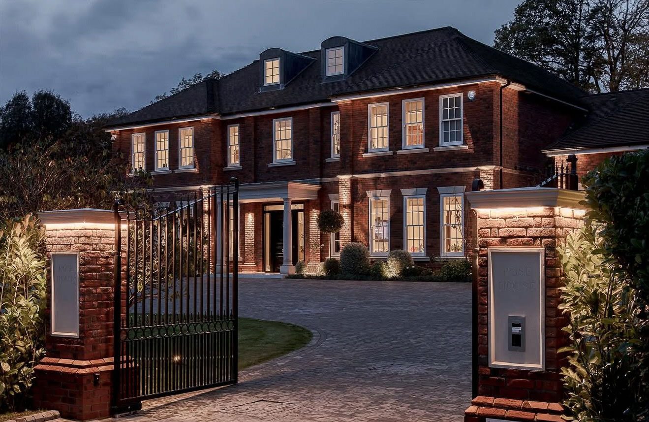 Front view of a 10,000 sq ft new build property in Kingswood, Surrey, showcasing red brick architecture, wrought iron gates and warm exterior lighting