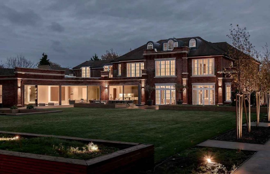 Rear elevation of a 10,000 sq ft luxury new build house in Kingswood, Surrey, with large sliding glass doors, landscaped gardens and evening architectural lighting