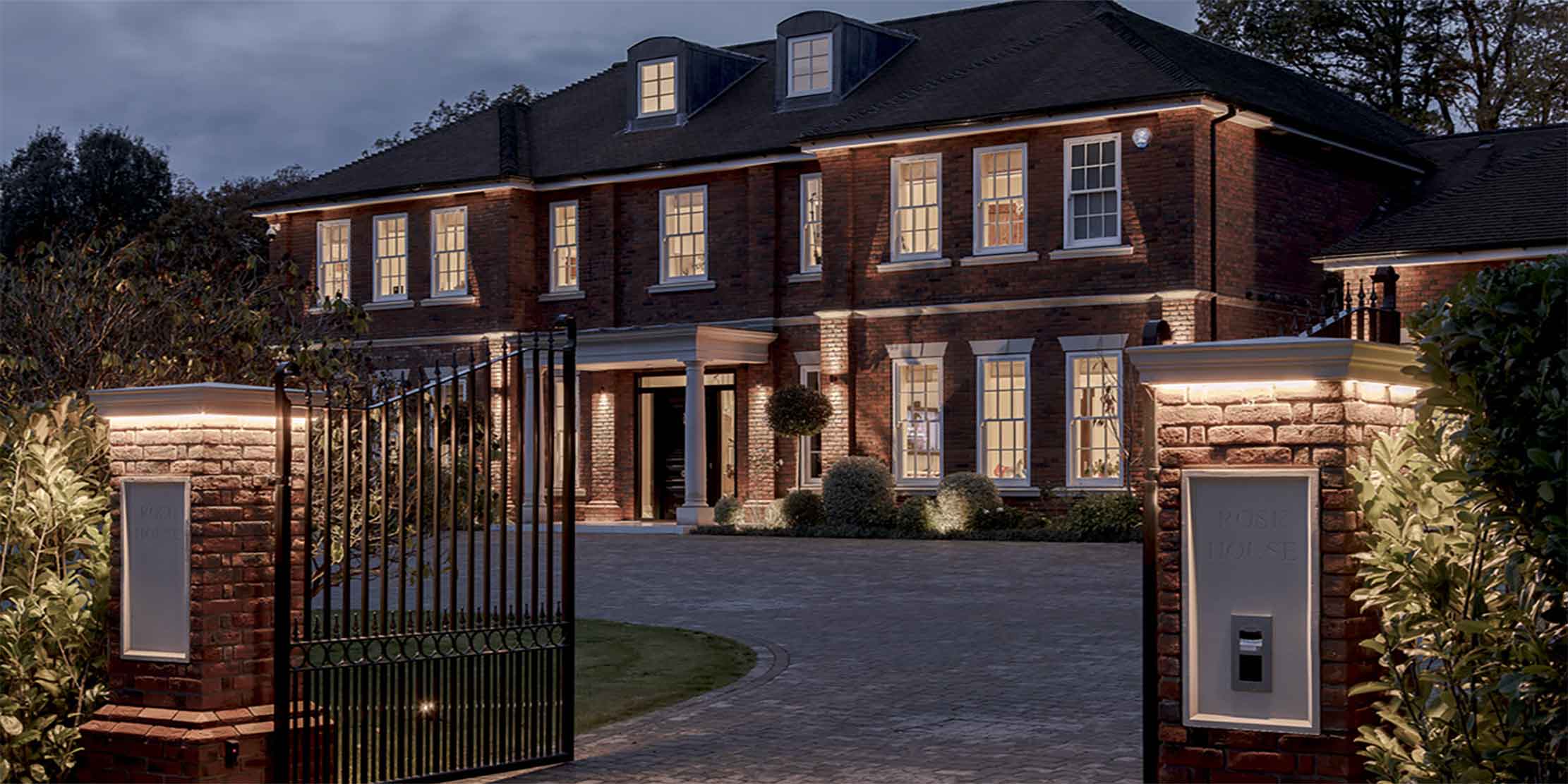 Luxury Georgian-style brick mansion exterior at dusk with bespoke architectural lighting, gated entrance, landscaped driveway, and premium residential design detailing.