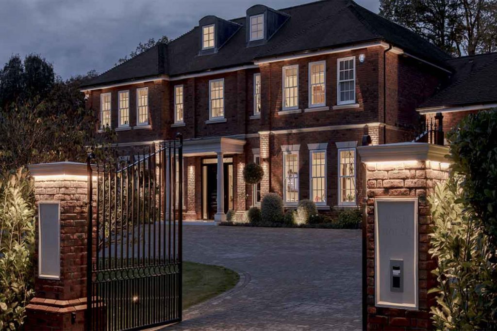 Luxury Georgian-style brick mansion exterior at dusk with bespoke architectural lighting, gated entrance, landscaped driveway, and premium residential design detailing.