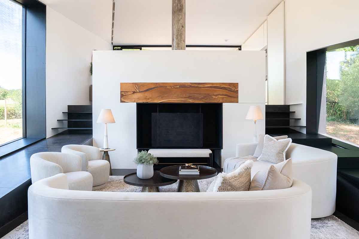 Modern minimalist living room with curved white sofa, sculptural armchairs, contemporary fireplace feature wall, black floating shelves, and floor-to-ceiling windows.