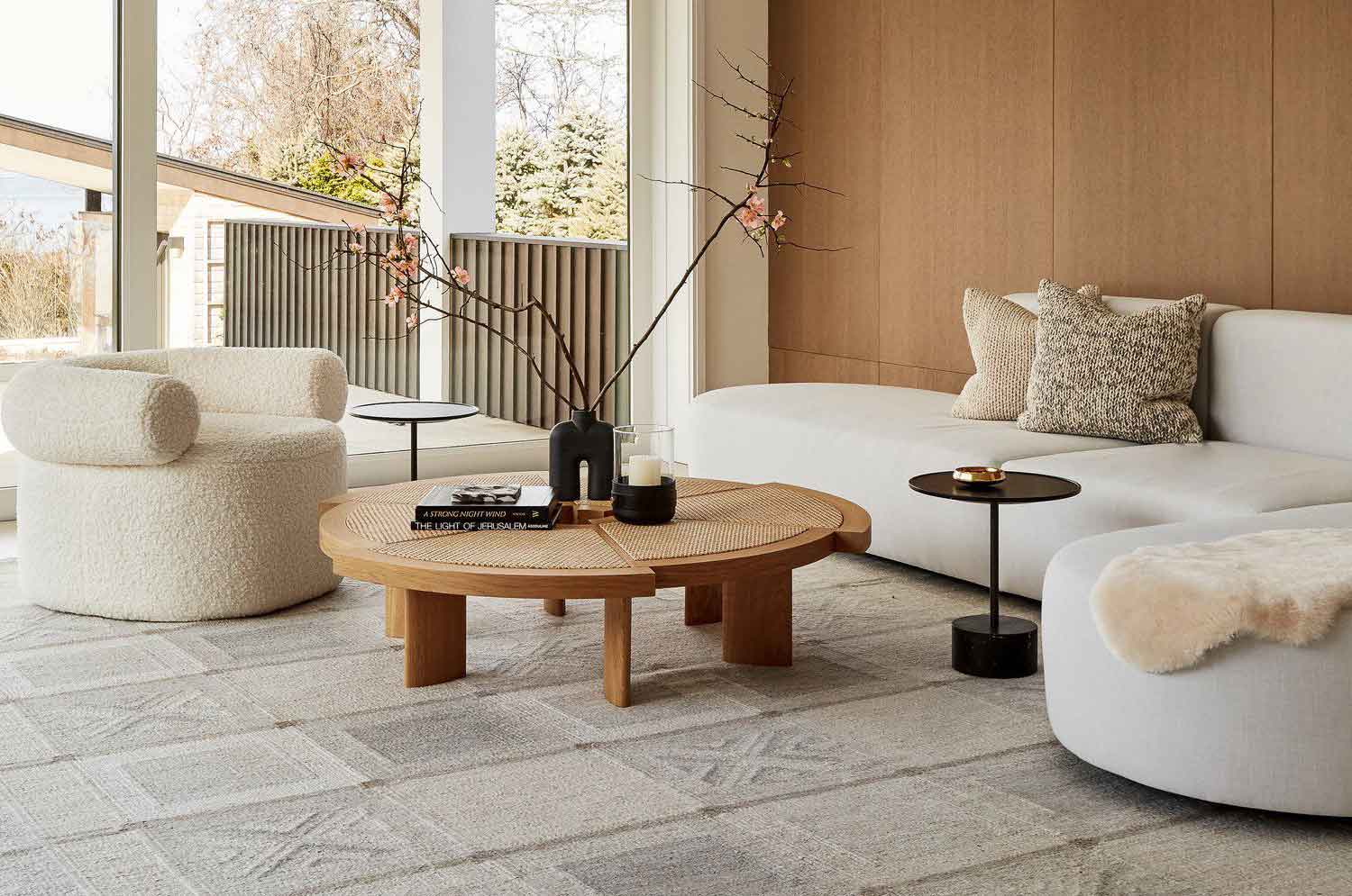 Organic modern living room with curved bouclé armchair, neutral sectional sofa, round wood coffee table, textured rug, and floor-to-ceiling windows with natural light.