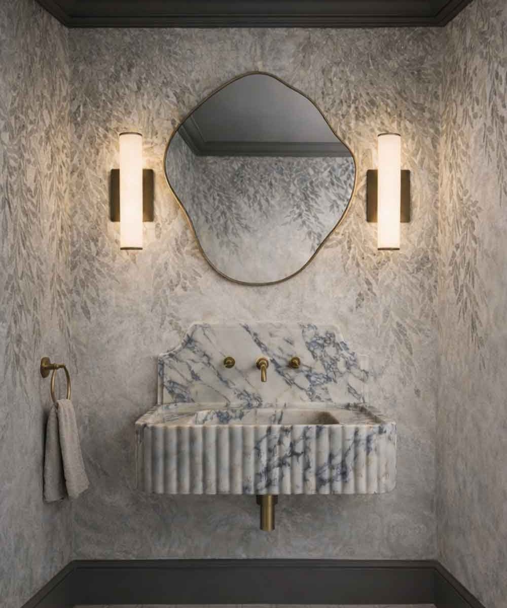 Luxury cloakroom with a sculptural marble sink, organic brass-framed mirror, warm wall sconces, textured wallpaper, and dark trim.