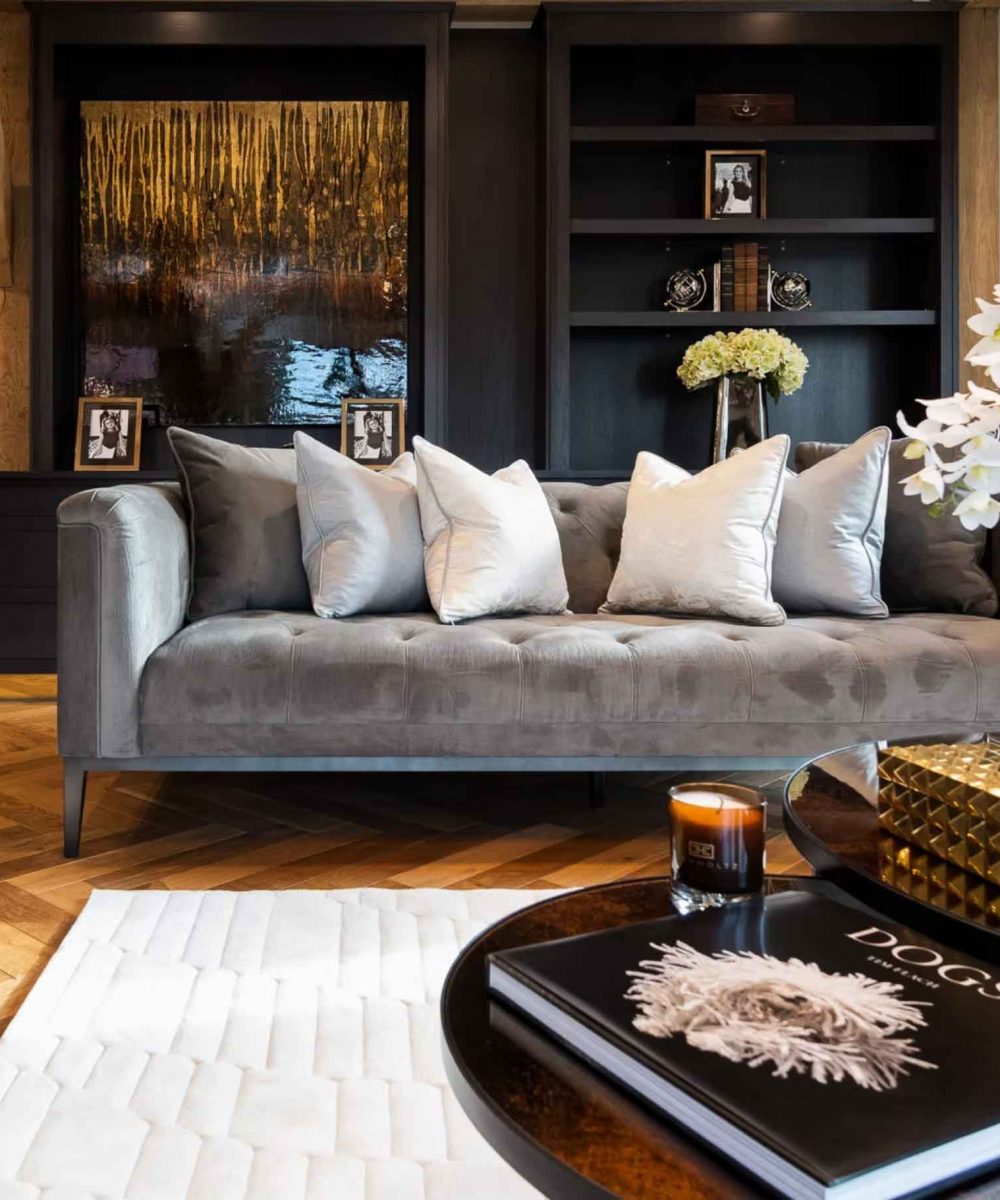 Luxury living room interior with grey velvet sofa, parquet flooring, dark built-in shelving, and elegant coffee table styling.